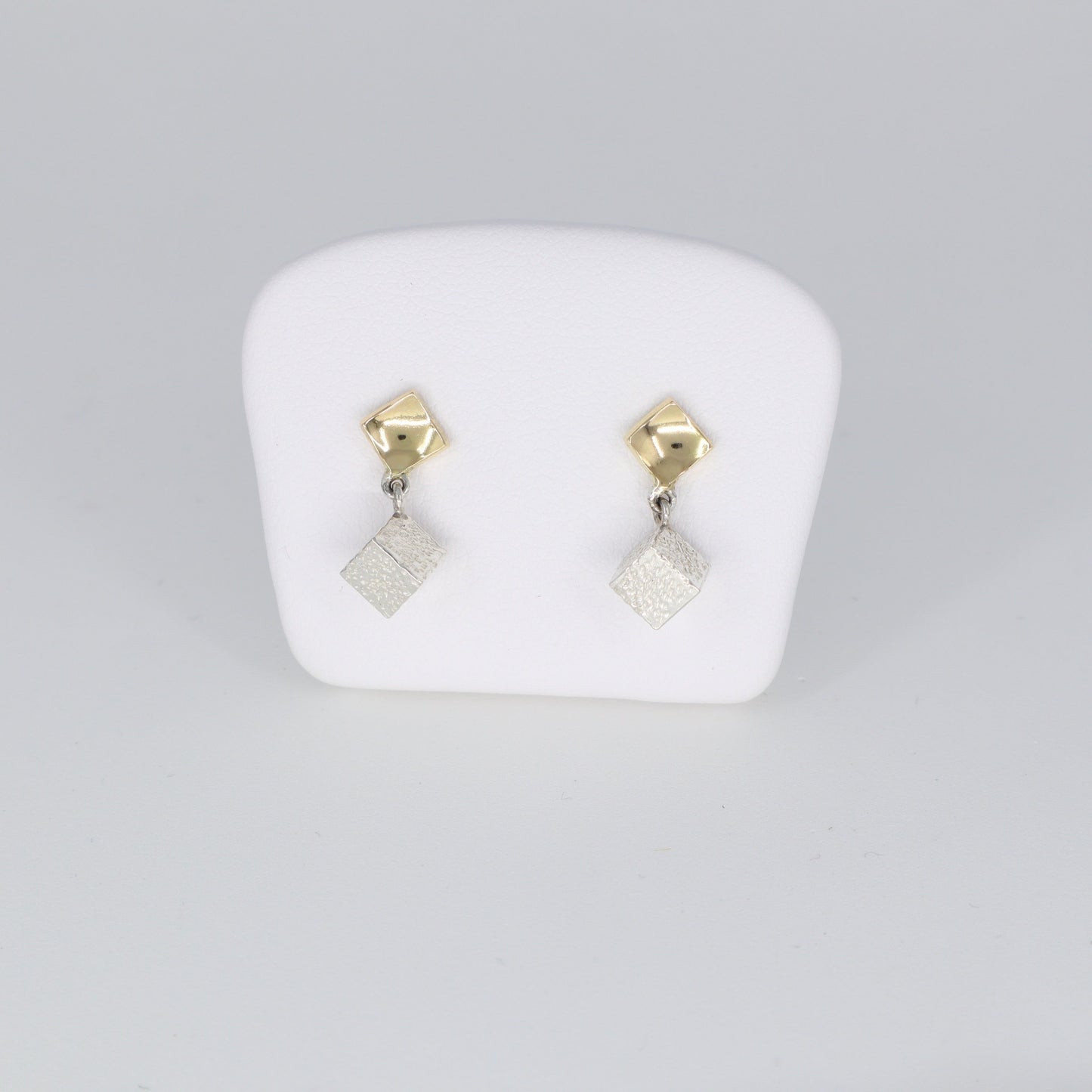 Voici une alternative pour la description de vos boucles d'oreilles :  "Boucles d'oreilles composées d'un carré en or 18 carats poli porté au lobe d'oreille, accompagné d'un cube en argent 925 martelé suspendu. Leur longueur totale est de 1,8 cm et leur largeur est de 0,5 cm. Un mariage élégant entre l'éclat de l'or et la texture du martelage en argent, ces boucles d'oreilles ajoutent une touche de sophistication à votre tenue quotidienne."