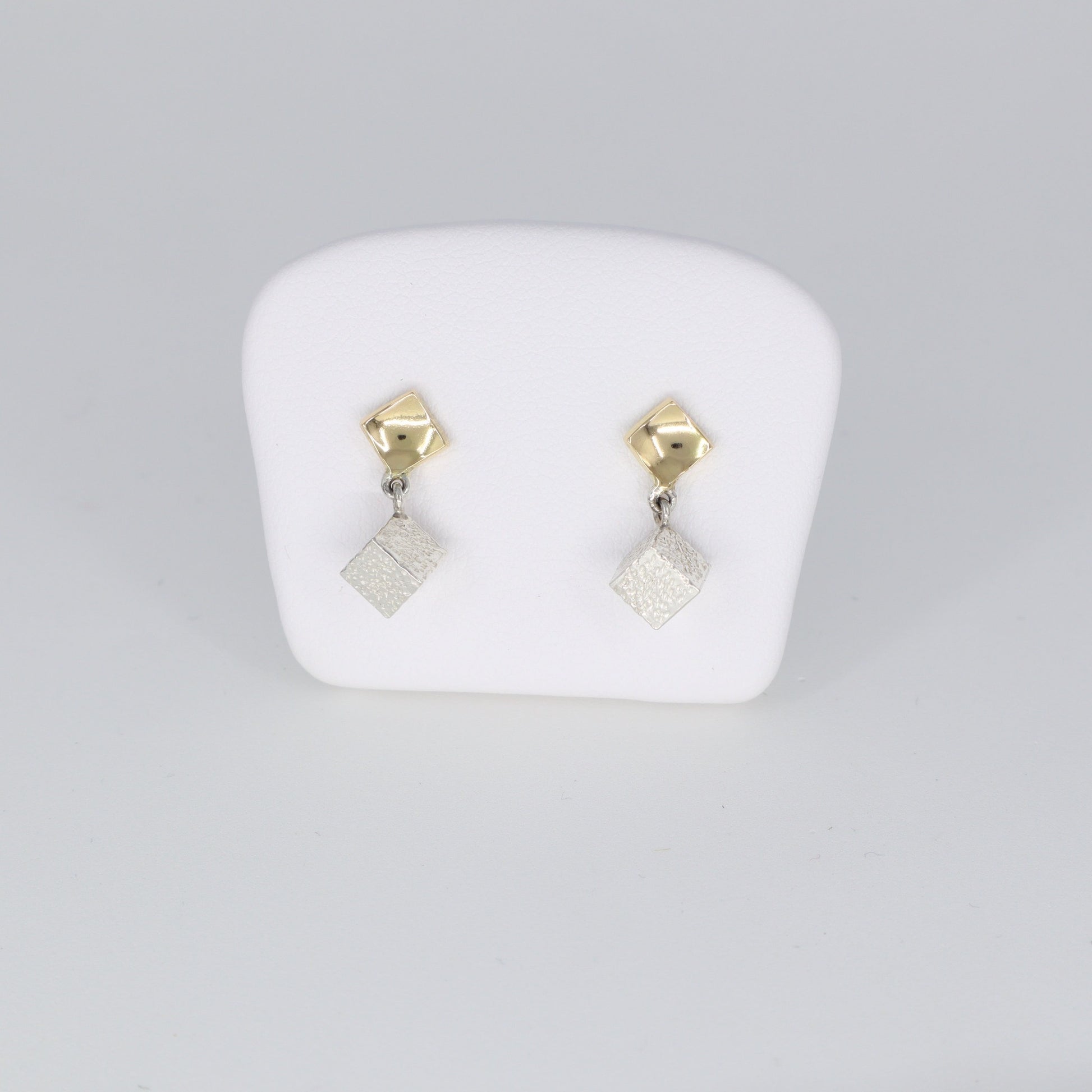 Voici une alternative pour la description de vos boucles d'oreilles :  "Boucles d'oreilles composées d'un carré en or 18 carats poli porté au lobe d'oreille, accompagné d'un cube en argent 925 martelé suspendu. Leur longueur totale est de 1,8 cm et leur largeur est de 0,5 cm. Un mariage élégant entre l'éclat de l'or et la texture du martelage en argent, ces boucles d'oreilles ajoutent une touche de sophistication à votre tenue quotidienne."