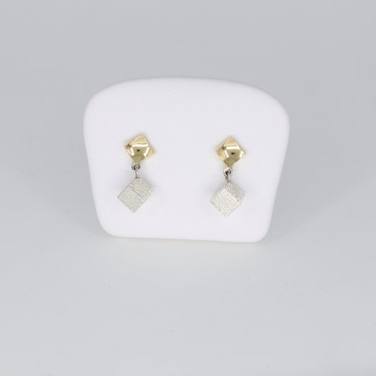 Voici une alternative pour la description de vos boucles d'oreilles :  "Boucles d'oreilles composées d'un carré en or 18 carats poli porté au lobe d'oreille, accompagné d'un cube en argent 925 martelé suspendu. Leur longueur totale est de 1,8 cm et leur largeur est de 0,5 cm. Un mariage élégant entre l'éclat de l'or et la texture du martelage en argent, ces boucles d'oreilles ajoutent une touche de sophistication à votre tenue quotidienne."