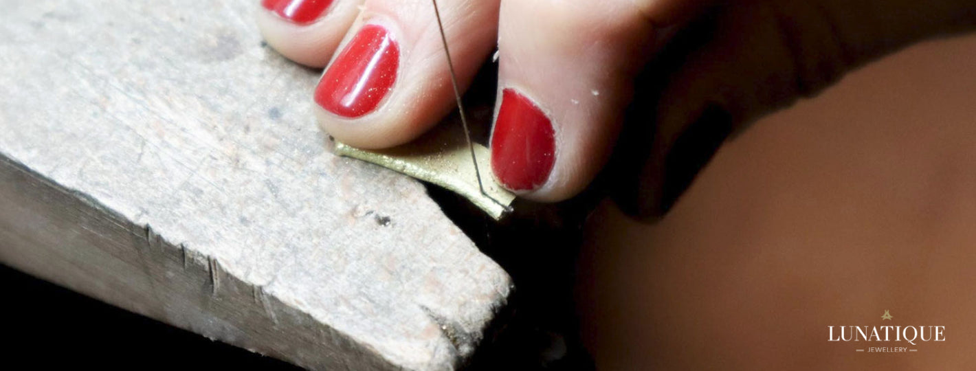 Trois doigts aux ongles courts, peints en rouge vif, tiennent fermement une plaque en or tandis qu'une fine lame de scie de bijoutier est en train de découper méticuleusement le métal précieux. Cette image captivante illustre le travail artisanal minutieux et la précision nécessaires dans la création de bijoux uniques et raffinés.